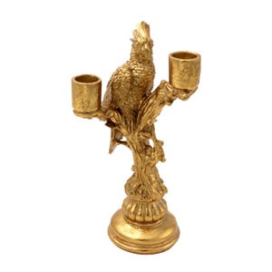 May include: A gold-colored parrot-shaped candle holder. The bird is perched on a tree branch, with two cylindrical holders for candles. The base is round and ornate. The candle holder is made of metal and has a detailed, textured surface.