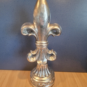 Könnte beinhalten: Eine silberfarbene Fleur-de-Lis-Skulptur. Das dekorative Stück hat ein detailliertes Design mit einer spitzen Spitze, geschwungenen Blütenblättern und einem dekorativen Sockel. Die Skulptur besteht aus einem metallischen Material und eignet sich für die Inneneinrichtung.