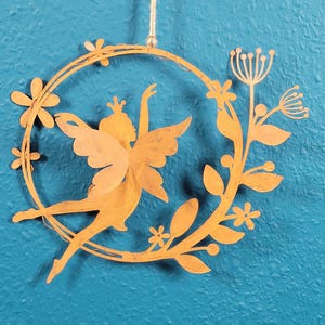 Puede incluir: Silueta de hada de metal dorado dentro de una corona floral, suspendida por una cuerda de hilo natural. El hada tiene los brazos extendidos y lleva una corona. La corona incluye flores y hojas, todo sobre un fondo turquesa.