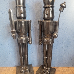 Puede incluir: Dos figuras de cascanueces de metal negro. Uno sostiene una espada, el otro un cetro. Ambos llevan sombreros altos y se paran sobre bases cuadradas. Las figuras están pintadas con un acabado negro brillante y son artículos decorativos.