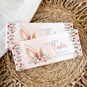 Pode incluir: Dois embrulhos de barra de chocolate de Páscoa rosa e branca com orelhas de coelho e desenhos florais. O texto na embalagem diz "Feliz Páscoa" e "Muito amor, A família Coelho".