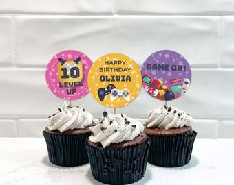 May include: Three chocolate cupcakes in black liners topped with white frosting and circular paper toppers. The toppers feature video game graphics, the text 10 LEVEL UP, HAPPY BIRTHDAY OLIVIA, and GAME ON, with illustrations of game controllers.