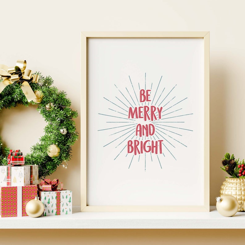 May include: A white framed print with the text "Be Merry and Bright" in red and blue lettering with a blue sunburst design behind the text. The print is on a white shelf with a Christmas wreath, ornaments, and wrapped gifts.