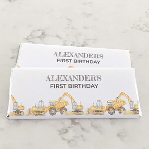 Pode incluir: Duas embalagens brancas de barras de chocolate com interiores de folha de prata. As embalagens apresentam uma fileira de veículos de construção amarelos e o texto "ALEXANDER'S FIRST BIRTHDAY" em cinza.