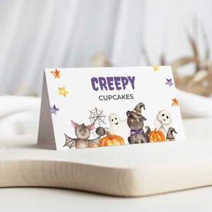 May include: A white card with the text "Creepy Cupcakes" in purple letters. The card is decorated with a black cat wearing a witch's hat, a ghost, a bat, a spider, and pumpkins.