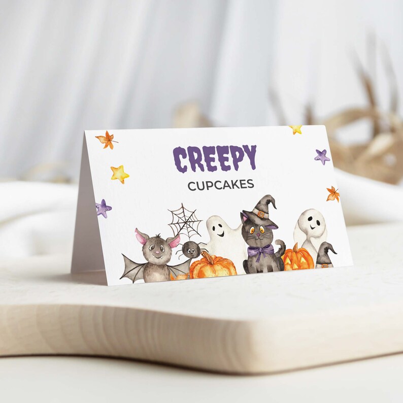 May include: A white card with the text "Creepy Cupcakes" in purple letters. The card is decorated with a black cat wearing a witch's hat, a ghost, a bat, a spider, and pumpkins.