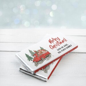 May include: Two rectangular chocolate bars wrapped in white paper featuring a red vintage truck carrying Christmas trees and gifts. The text reads Merry Christmas! Lots of love, The Claus Family in red and black script and block lettering.