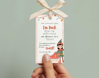 May include: A rectangular gift tag with a cream-colored ribbon at the top. The tag features a cartoon elf, a red sleigh, and text that reads: Dear Emma and Sophie, I'm Back FROM THE NORTH POLE AND I BROUGHT YOU A TREAT! I'll be reporting to Santa each day so remember to be extra sweet! Love your Elf, Holly.