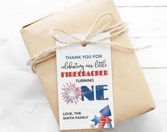 Puede incluir: Una etiqueta de regalo rectangular blanca atada con cordel a un paquete envuelto en papel marrón. La etiqueta presenta texto en rojo, blanco y azul: THANK YOU FOR celebrating our little FIRECRACKER TURNING ONE, con un gráfico de fuegos artificiales y THE SMITH FAMILY.