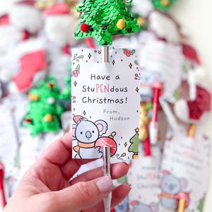 May include: A silver pen topped with a green sequined Christmas tree featuring small red and yellow pom-poms. Attached is a white card with a koala illustration and the text: Have a StuPENdous Christmas! From, Hudson. The pen is metallic silver.