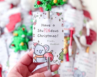 May include: A silver pen topped with a green sequined Christmas tree featuring small red and yellow pom-poms. Attached is a white card with a koala illustration and the text: Have a StuPENdous Christmas! From, Hudson. The pen is metallic silver.