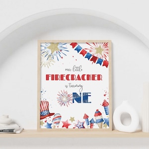Puede incluir: Una lámina rectangular en un marco de madera clara con el texto 'our little FIRECRACKER is turning ONE' en rojo y azul. El diseño incluye ilustraciones en acuarela de estrellas doradas, fuegos artificiales, un sombrero de copa y cohetes.