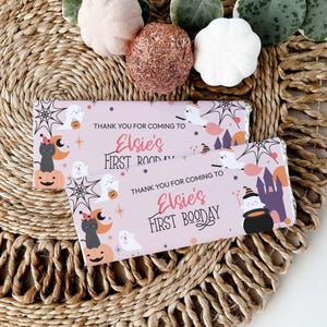 Puede incluir: Dos envoltorios rosas de barras de caramelo con gráficos temáticos de Halloween. Los envoltorios presentan fantasmas, calabazas y un gato negro. El texto dice "Thank you for coming to Elsie's First Booday."