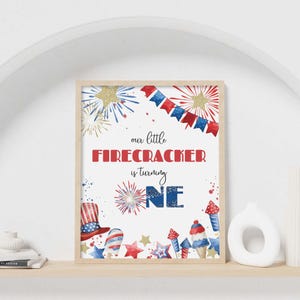 May include: A framed print with a patriotic theme. The text reads "our little FIRECRACKER is turning ONE" in red and blue. The print features watercolor illustrations of fireworks, stars, and a festive hat, in red, white, and blue.