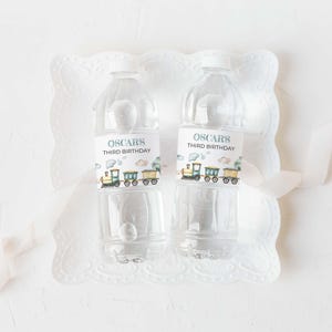 Könnte beinhalten: Zwei transparente Plastikwasserflaschen mit weißen Deckeln, jeweils mit der Aufschrift "OSCAR'S THIRD BIRTHDAY". Die Etiketten zeigen eine Zugillustration in Grün, Gelb und Blau. Die Flaschen stehen auf einem weißen, dekorativen Tablett mit gewelltem Rand.