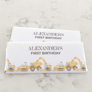 May include: Two rectangular chocolate bar wrappers with a white background. The text reads ALEXANDER'S FIRST BIRTHDAY in black lettering. Below the text, a repeating pattern of yellow construction excavators and dump trucks is printed.