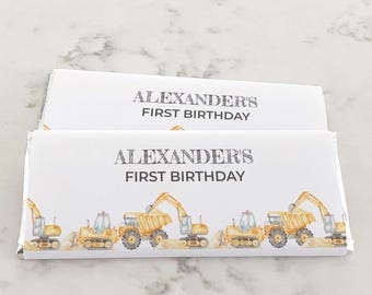 May include: Two rectangular chocolate bar wrappers with a white background. The text reads ALEXANDER'S FIRST BIRTHDAY in black lettering. Below the text, a repeating pattern of yellow construction excavators and dump trucks is printed.