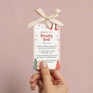 May include: A rectangular tag with the text "MAGICAL ELF Recovery Dust Instructions" and a cream-coloured bow. The tag has a pink background with snowflakes and Christmas tree illustrations. The tag is held by a hand.