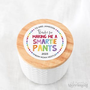 Può includere: Un barattolo in ceramica bianca con coperchio in legno chiaro dotato di un adesivo circolare con la scritta TO MRS JAMESON Thanks for MAKING ME A SMARTIE PANTS 2023 FROM NOAH NESTLE in lettere colorate e decorate con un bordo a pois.