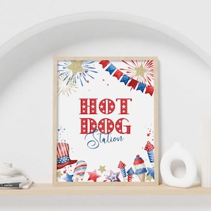 May include: A rectangular print in a light wood frame featuring the red text HOT DOG and the blue script text Station. The design includes watercolor illustrations of red, white, and blue fireworks, stars, bunting, and patriotic themed treats.