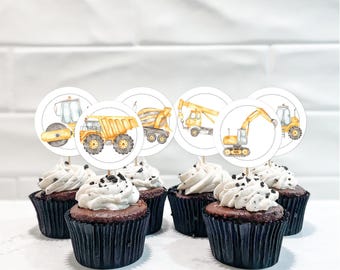 Op de afbeelding: Een set chocoladecupcakes in zwarte vormpjes met witte topping en koekkruimels, elk voorzien van een ronde papieren topper met een aquarelillustratie van een geel bouwvoertuig zoals een graafmachine of kiepwagen.