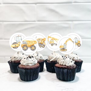 May include: Six chocolate cupcakes with white frosting and dark sprinkles, topped with construction vehicle-themed cupcake toppers. The toppers feature illustrations of a road roller, dump truck, crane, excavator, and other construction vehicles.
