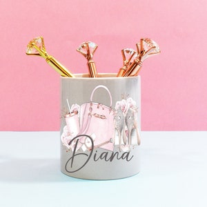 Può includere: Un portapenne in ceramica grigia con una borsa a mano rosa, tacchi alti e il nome "Diana" stampato su di esso. Il portapenne contiene penne dorate con punte a forma di diamante.