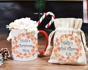 May include: A white ceramic mug with a red handle and a gingerbread design. The mug says "Holly's Christmas Mug". A white burlap bag with a gingerbread design says "Holly's Hot Choc Kit".