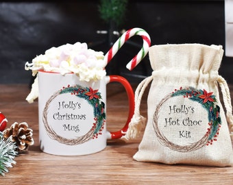 May include: A white ceramic mug with a red handle and a Christmas wreath design. The mug says "Holly's Christmas Mug". A white canvas bag with a drawstring closure says "Holly's Hot Choc Kit".