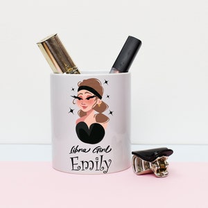 May include: White ceramic makeup brush holder with a cartoon illustration of a woman with brown hair and black eyeliner. The text "Libra Girl Emily" is printed on the holder.