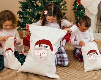 May include: Three personalized Santa sack gift bags with red and white trim. The bags are decorated with a cartoon Santa Claus face and have the names "Emma", "Lila", and "Thomas" printed on them.