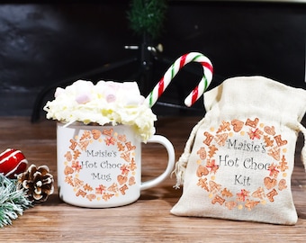 May include: A white mug with a gingerbread design and the text "Maisie's Hot Choc Mug" is filled with hot chocolate, whipped cream, and marshmallows. A burlap bag with the text "Maisie's Hot Choc Kit" and a gingerbread design is next to the mug. A candy cane is leaning against the mug.