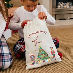 May include: A white canvas gift bag with a red drawstring. The bag has a watercolor illustration of a Christmas tree, a nutcracker, and a mouse in a nutcracker costume. The text "Special delivery for Thomas" is printed in red on the bag.