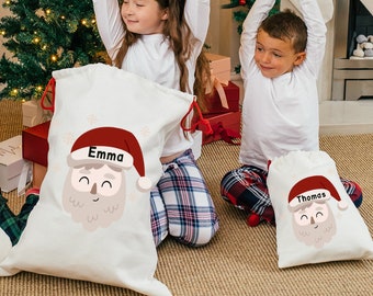 May include: Two personalized Christmas gift bags with Santa Claus faces. The bags are white with red trim and have the names "Emma" and "Thomas" printed on them.