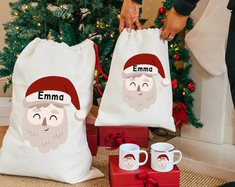 May include: Two white canvas Santa sacks with red trim and a cartoon Santa face with the name "Emma" on each. The sacks are sitting on a red box with two white mugs with Santa faces on them.