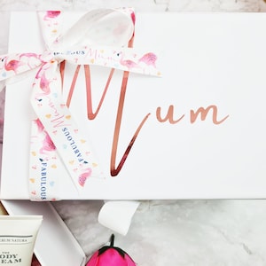 K&ouml;nnte beinhalten: Wei&szlig;e Geschenkbox mit einem rosa Band und einem Flamingo-Druck. Die Box ist mit dem Wort "Mum" in ros&eacute;goldenen Lettern verziert.