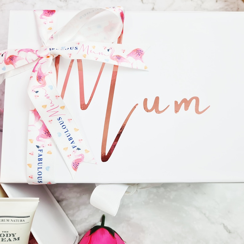 K&ouml;nnte beinhalten: Wei&szlig;e Geschenkbox mit einem rosa Band und einem Flamingo-Druck. Die Box ist mit dem Wort "Mum" in ros&eacute;goldenen Lettern verziert.
