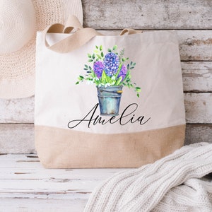 May include: A white canvas tote bag with a brown burlap bottom. The bag features a watercolor illustration of a bucket of purple hyacinths and the name "Amelia" in cursive.