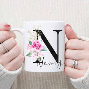 K&ouml;nnte beinhalten: Wei&szlig;e Keramiktasse mit einem schwarzen Buchstaben N und einem rosafarbenen Blumendesign. Die Tasse sagt "Nanny" unter dem Buchstaben.