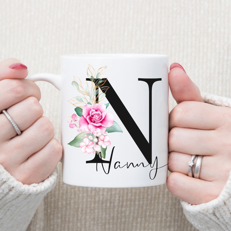 K&ouml;nnte beinhalten: Wei&szlig;e Keramiktasse mit einem schwarzen Buchstaben N und einem rosafarbenen Blumendesign. Die Tasse sagt "Nanny" unter dem Buchstaben.