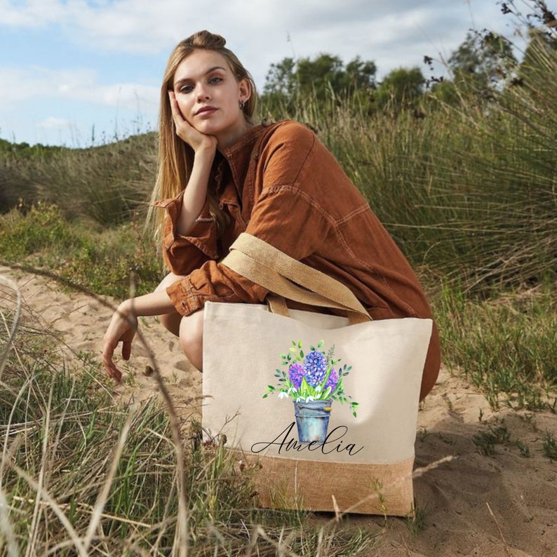 May include: A personalized tote bag with a floral design and the name "Amelia" printed on the front. The bag is made of natural canvas and has a jute trim.