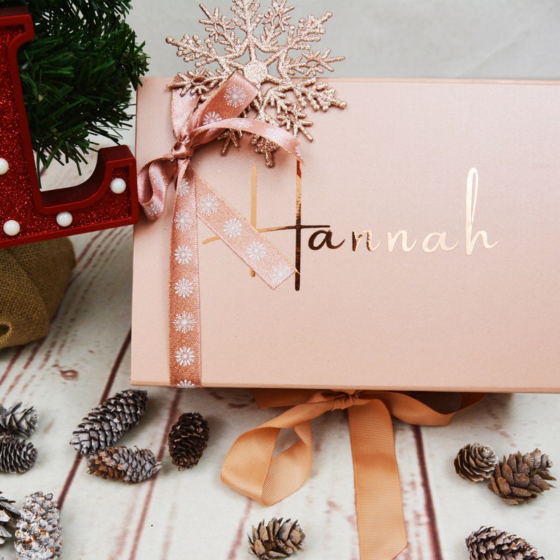 May include: A pink gift box with a gold ribbon and a snowflake decoration. The box is tied with a brown ribbon and the name "Hannah" is written in gold on the front.