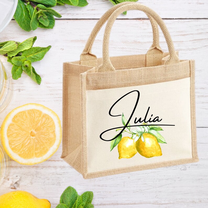 K&ouml;nnte beinhalten: Eine kleine, naturfarbene Jute-Tragetasche mit einem wei&szlig;en Canvas-Einsatz. Der Einsatz zeigt eine Aquarell-Illustration von Zitronen und einen handgeschriebenen Namen "Julia".