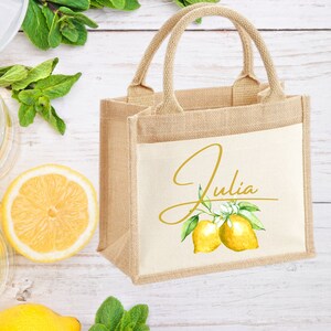 K&ouml;nnte beinhalten: Eine kleine, naturfarbene Jute-Tragetasche mit einem wei&szlig;en Canvas-Einsatz. Der Einsatz zeigt eine goldene Schriftart in Schreibschrift, die "Julia" mit einem Zitronenzweig und zwei Zitronen im Aquarell-Stil sagt.