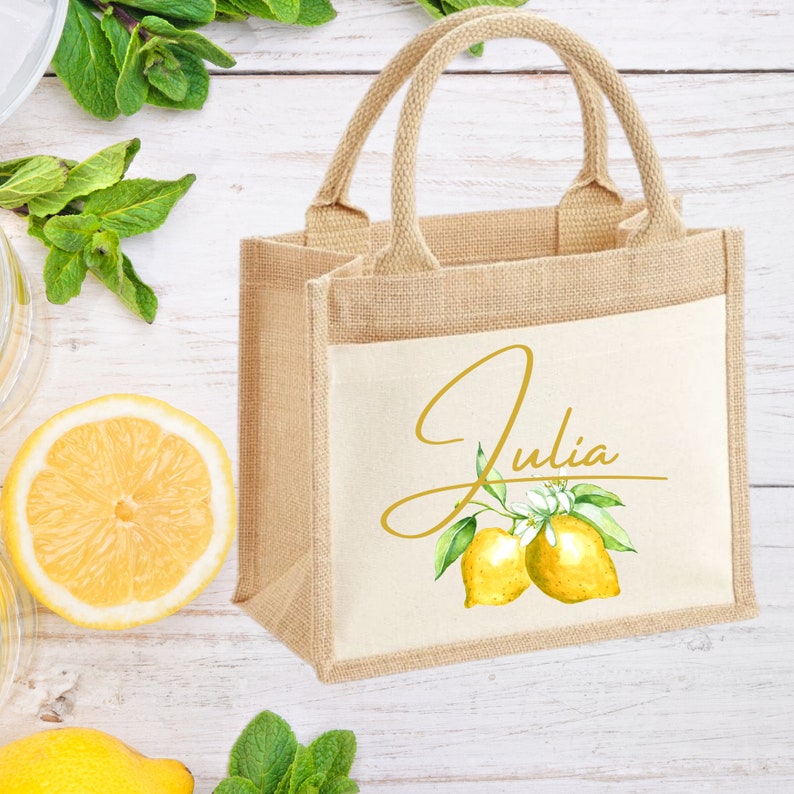 K&ouml;nnte beinhalten: Eine kleine, naturfarbene Jute-Tragetasche mit einem wei&szlig;en Canvas-Einsatz. Der Einsatz zeigt eine goldene Schriftart in Schreibschrift, die "Julia" mit einem Zitronenzweig und zwei Zitronen im Aquarell-Stil sagt.