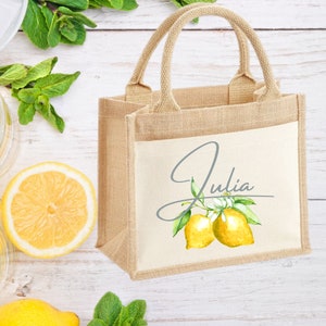 K&ouml;nnte beinhalten: Eine kleine, naturfarbene Jute-Tragetasche mit einem wei&szlig;en Canvas-Einsatz. Der Canvas-Einsatz zeigt eine Aquarell-Illustration von Zitronen und einen handschriftlichen Namen, "Julia".