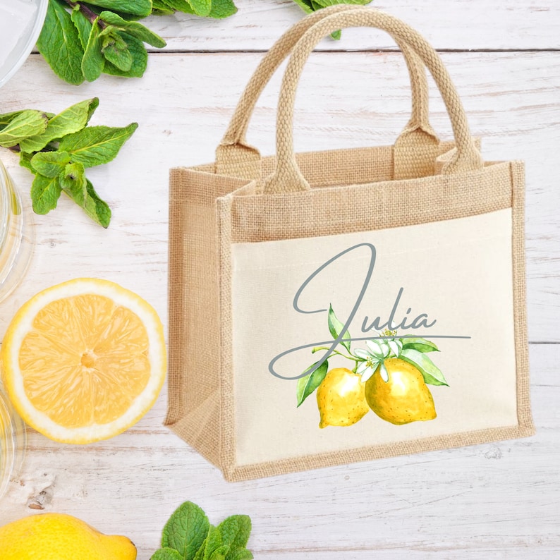 K&ouml;nnte beinhalten: Eine kleine, naturfarbene Jute-Tragetasche mit einem wei&szlig;en Canvas-Einsatz. Der Canvas-Einsatz zeigt eine Aquarell-Illustration von Zitronen und einen handschriftlichen Namen, "Julia".