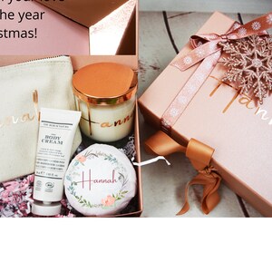 May include: A pink gift box with a gold snowflake and ribbon, a white gift bag with the name "Hannah" written on it, a white body cream tube, and a glass candle with a gold lid and the name "Hannah" written on it.