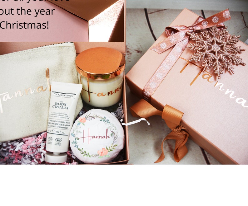 May include: A pink gift box with a gold snowflake and ribbon, a white gift bag with the name "Hannah" written on it, a white body cream tube, and a glass candle with a gold lid and the name "Hannah" written on it.