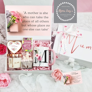 K&ouml;nnte beinhalten: Eine wei&szlig;e Geschenkbox gef&uuml;llt mit einer rosa Tasse mit floralem Design und dem Buchstaben "M", einer herzf&ouml;rmigen Box mit dem Wort "Mama" darauf, einer Kerze mit dem Wort "Mama" darauf und einer Schachtel mit Hei&szlig;getr&auml;nkemischung. Die Box ist mit rosa Seidenpapier und rosa Blumen verziert. Die Box enth&auml;lt auch eine Karte mit dem Text "Mama" und einer Dankesbotschaft.